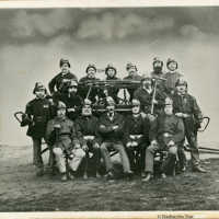 Schwarz-weiß Foto: In drei Reihen stehen und sitzen die 15 Feuerwehrmänner von 1875. Sie tragen Uniform und halten Werkzeug in den Händen. 