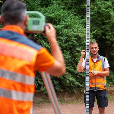 Junger Mann hält Teleskopnivellierlatte, während ein Kollege Messungen anstellt