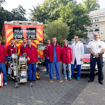 Das ECMO-Team des Brüderkrankenhauses Trier mit Thomas Reinholz, dem Leiter des Rettungsdienstes der Feuerwehr (ganz rechts) und Ralf Cordel von der Berufsfeuerwehr (ganz links) vor dem Intensivtransportwagen und dem Begleitfahrzeug.