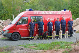 Die Mitglieder stehen gemeinsam mit ihren Hunden vor dem Wagen der Rettungshundestaffel.