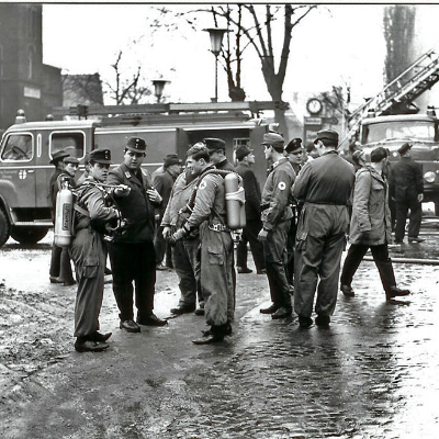 Schwarz-weiß Foto: Zahlreiche Feuerwehrleute und Passanten stehen auf der Ehranger Straße und unterhalten sich. Im Hintergrund stehen zwei Feuerwehrfahrzeuge und eine ausgefahrene Leiter.
