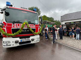 Im Vordergrund des Fotos ist die geschmückte Front eines Feuerwehrautos zu sehen. Im Hintergrund ein Pfarrer, der es einsegnet.