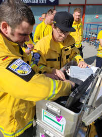 Auf dem Bild sind zwei Helfer in gelben Einsatzjacken zu sehen, die technische Ausrüstung in Metallkisten inspizieren.  