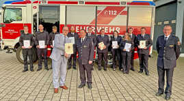 Feuerwehrdezernent Ralf Britten übergibt Wehrführer Bernd Becker seine Auszeichnung. Im Hintergrund stehen die weiteren ausgezeichneten Mitglieder der Freiwilligen Feuerwehr Irsch vor einem Feuerwehrfahrzeug. 