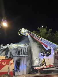 Ein Drehleiterfahrzeug der Feuerwehr löscht ein Haus mit Schaum. Es ist Nacht, eine Straßenlaterne scheint.