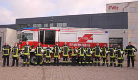 Die 13 neuen freiwilligen Feuerwehrleute stehen gemeinsam mit ihren beiden Ausbildern in Feuerwehrausrüstung vor einem Feuerwehrfahrzeug auf dem Hof der Feuerwache 2 in Ehrang.