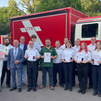 Eine größere Gruppe von Menschen steht vor einem Feuerwehrfahrzeug. Viele präsentieren ihre Urkunden.