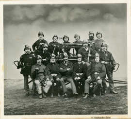 Schwarz-weiß Foto: In drei Reihen stehen und sitzen die 15 Feuerwehrmänner von 1875. Sie tragen Uniform und halten Werkzeug in den Händen. 