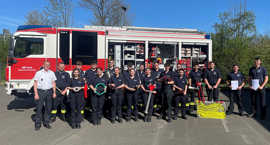 Junge männliche bund weibliche Feuerwehrleute stehen in zwei Reihen vor einem Löschfahrzeug und halten Feuerwehr-Gerätschaften und -Zubehöt in ihren Händen