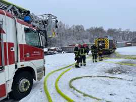 Auf einem verschneiten Firmengelände stehen zwei Einsatzfahrzeuge der Feuerwehr. Feuerwehrleute in Schutzkleidung haben gelbe Schläuche verlegt.