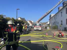 Feuerwehrleute in Einsatzmontur und ein Fahrzeug mit ausgefahrener Drehleiter stehen in einer Straße, auf der gelbe Schläuche verlegt sind.