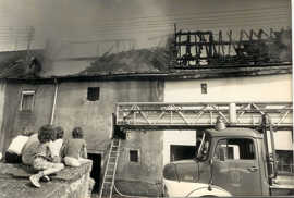 Ein Drehleiterfahrzeug steht vor den beiden Häusern mit abgebrannten Dachstühlen. Fünf Kinder sitzen auf einer Mauer und beobachten die Szene. 